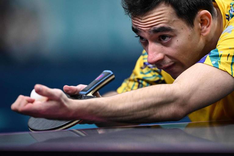 Um jogador de tênis de mesa está se preparando para fazer um saque. Ele está inclinado sobre a mesa, com uma expressão concentrada. O jogador usa uma camiseta amarela e segura uma raquete na mão direita, enquanto a bola está posicionada na mão esquerda.