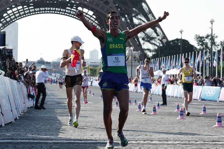 A imagem mostra um atleta levantando os braços em sinal de vitória enquanto cruza a linha de chegada de uma corrida. Ele está vestido com um uniforme verde e amarelo, representando o Brasil. Ao fundo, há outros corredores e uma estrutura arquitetônica, possivelmente a Torre Eiffel, com bandeiras ao redor.