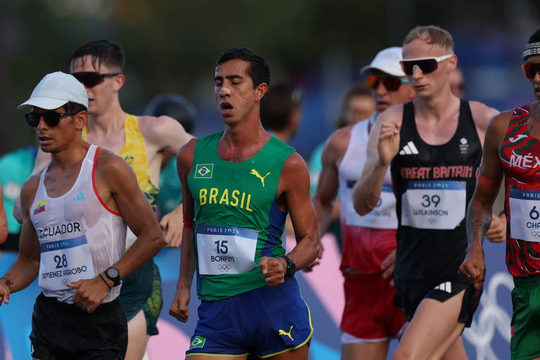 A imagem mostra um grupo de atletas competindo em uma corrida de atletismo. Entre eles, um atleta vestido com uma camiseta verde e amarela representa o Brasil. Os outros atletas estão usando uniformes de diferentes países. O cenário é ao ar livre, com um fundo desfocado que sugere um evento esportivo.