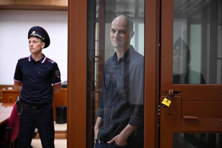 A imagem mostra um homem com cabeça raspada, sorrindo, dentro de uma cela de vidro em um tribunal. Ele está vestido com uma camisa xadrez e tem as mãos nos bolsos. Ao lado, um policial em uniforme, com um boné, está em pé, observando. O ambiente é de um tribunal, com paredes claras e móveis de madeira.