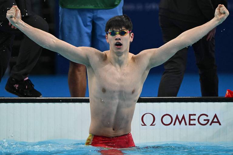 Um jovem atleta de natação, sem camisa e usando óculos de natação, está em uma piscina levantando os braços em celebração. Ele parece estar muito feliz e vitorioso. Ao fundo, é possível ver pessoas e parte da estrutura da piscina. Na borda da piscina, há um logotipo da marca Omega.