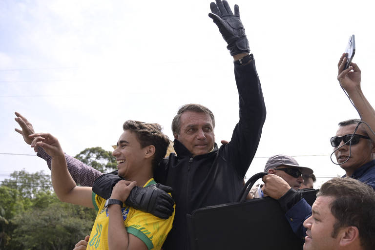 Na imagem, é possível ver Jair Bolsonaro, homem branco e de cabelos grisalhos abraçando o deputado federal Nikolas Ferreira, homem de cabelos castanhos. Bolsonaro veste jaqueta e luvas pretas. Nikolas uma camiseta da seleção brasileira de futebol. Eles estão em um local aberto