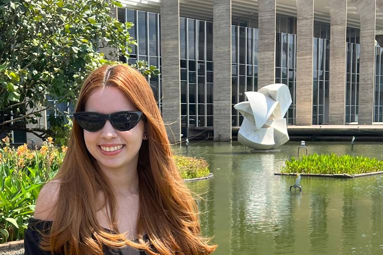 Uma mulher com cabelo longo e liso, usando óculos escuros, sorri em frente a um lago com plantas aquáticas. Ao fundo, há um edifício moderno com grandes janelas e uma escultura abstrata em forma de folhas. O ambiente é ensolarado e há vegetação ao redor.