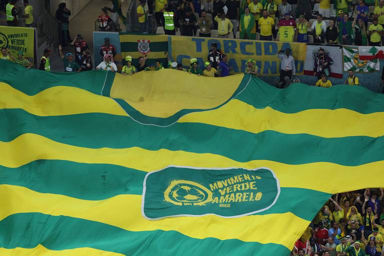 Fotografia tirada de cima mostra uma faixa gigante estendida pelo Movimento Verde Amarelo na arquibancada de um estádio durante partida de futebol do Grupo G da Copa do Mundo do Qatar 2022. A faixa tem o formato de uma camiseta e listras amarelas e verdes.