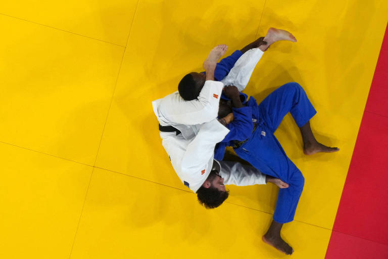 A imagem mostra dois lutadores de judô em uma competição. Um lutador está vestido com um kimono branco e o outro com um kimono azul. Eles estão em uma posição de grappling no tatame amarelo, com um lutador tentando aplicar uma técnica de controle sobre o outro.
