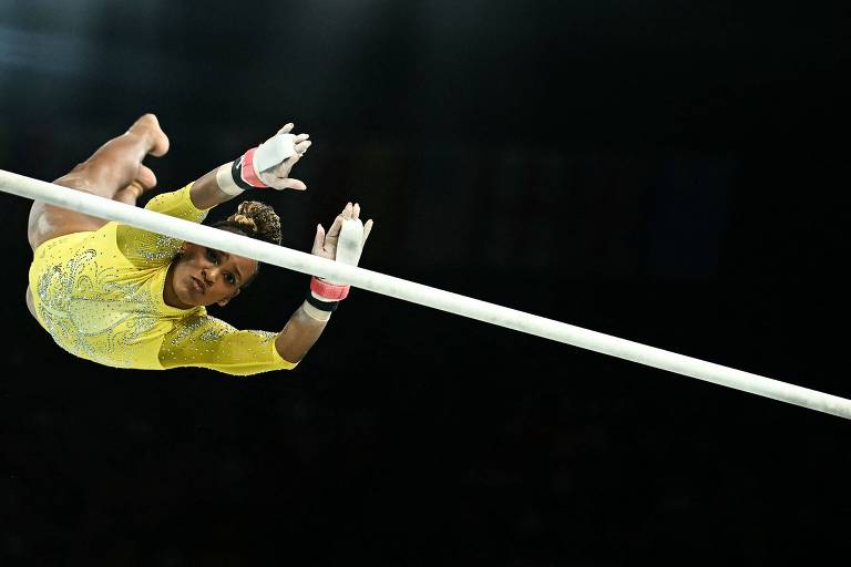 Um atleta está realizando um salto com vara, pendurado de cabeça para baixo, com as pernas para cima e as mãos segurando a vara. O atleta usa um traje amarelo e luvas, e o fundo é escuro, sugerindo um ambiente de competição.
