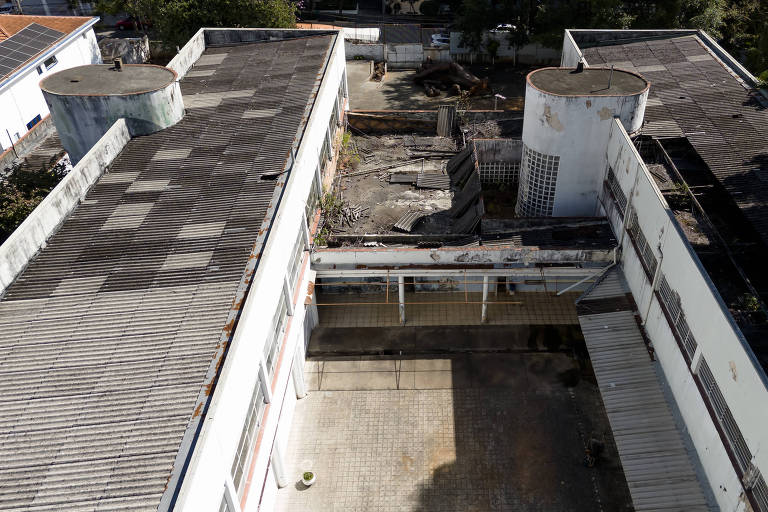 A imagem mostra uma vista aérea de um edifício abandonado com telhados de metal e estruturas deterioradas. O telhado é coberto por placas de metal, algumas com ferrugem visível. No centro, há uma área aberta com piso de ladrilhos, cercada por paredes brancas e janelas. Ao fundo, pode-se ver um espaço com entulho e vegetação ao redor. Dois tanques cilíndricos estão localizados em cada extremidade do edifício.