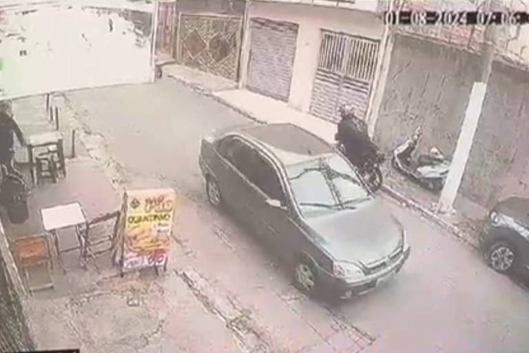 A imagem mostra uma rua vista de uma câmera de segurança. Um carro cinza passa pelo centro da rua, enquanto uma motocicleta, a do PM, está parada à direita
