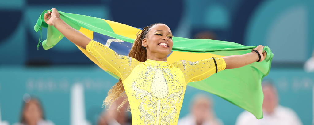 Uma ginasta está em uma apresentação, vestindo um traje amarelo e segurando uma bandeira verde e amarela do Brasil. Ela parece estar celebrando, com um sorriso no rosto, enquanto o público ao fundo observa.