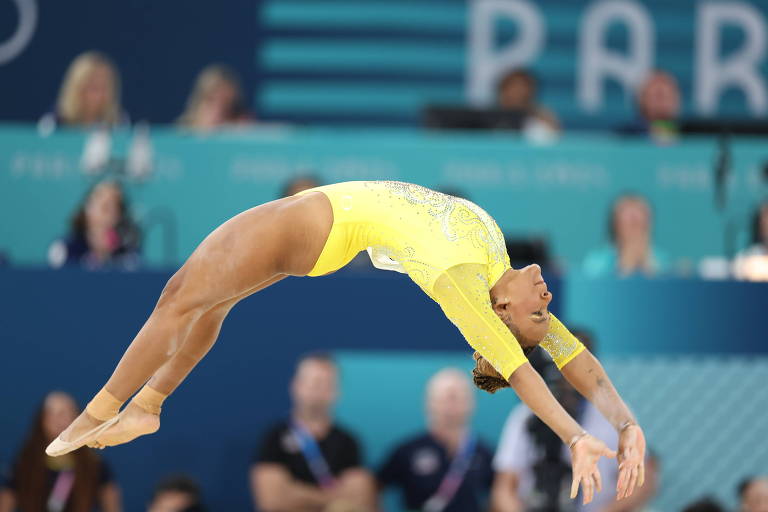 Uma ginasta está realizando um salto acrobático, com o corpo arqueado para trás. Ela veste um traje amarelo e está no ar, com os braços estendidos. Ao fundo, há uma plateia visível, com pessoas assistindo ao evento.
