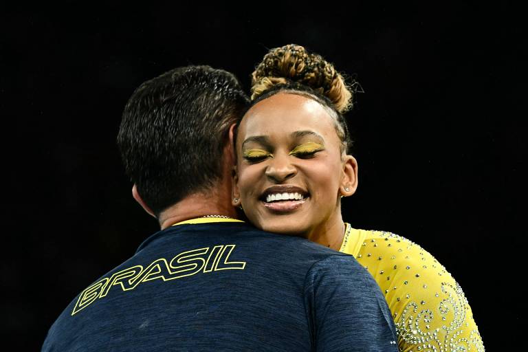 A ginasta Rebeca Andrade sorri ao abraçar seu treinador, que está de costas, depois de se apresentar nas barras paralelas assimétricas na final do individual geral em Paris-2024