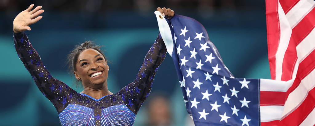 Uma ginasta está sorrindo e acenando enquanto segura a bandeira dos Estados Unidos. Ela está vestindo um traje de ginástica azul com detalhes em preto e está em um ambiente de competição, com um público ao fundo.