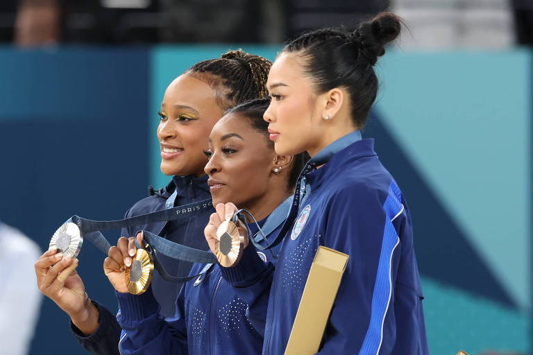 Três atletas estão no pódio, vestindo uniformes azuis e segurando medalhas. Elas estão sorrindo e posando para a câmera, enquanto uma multidão ao fundo aplaude. O ambiente parece ser uma cerimônia de premiação.