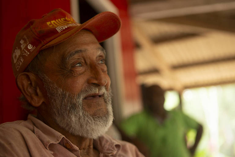 A imagem mostra um homem idoso com barba e cabelo grisalho, usando um boné vermelho. Ele está sorrindo e olhando para cima, com um fundo desfocado que sugere um ambiente interno. Ao fundo, é possível ver uma pessoa com uma camisa verde, mas não está claramente focada.