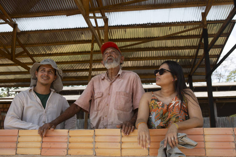 A imagem mostra três pessoas em um ambiente de construção. À esquerda, um jovem com um chapéu claro e camisa de manga longa observa com um sorriso. No centro, um homem idoso com barba e um chapéu vermelho está em pé, olhando para cima, com uma expressão pensativa. À direita, uma mulher com óculos escuros e uma blusa colorida está sentada, olhando para o homem idoso. Eles estão próximos a uma parede feita de tijolos de cor laranja, e o teto é de metal ondulado.