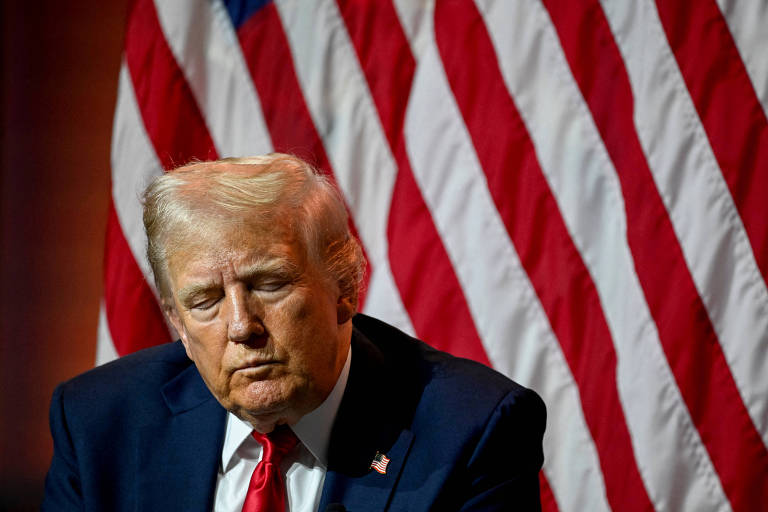 A imagem mostra um homem com cabelo loiro e terno escuro, sentado em frente a uma bandeira dos Estados Unidos, que tem listras vermelhas e brancas e um campo azul com estrelas. O homem parece pensativo e está olhando para baixo.