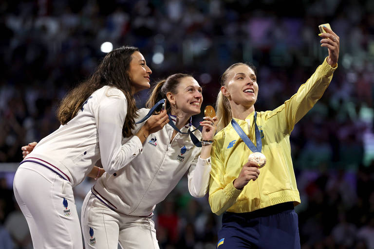 Três atletas estão no pódio, sorrindo e posando para uma selfie. A atleta à direita está segurando uma medalha de ouro, enquanto as outras duas atletas, uma à esquerda e outra ao centro, estão usando uniformes brancos e segurando medalhas de prata e bronze. O fundo mostra uma multidão em um evento esportivo.

