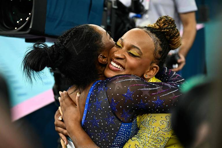 A imagem mostra duas mulheres se abraçando com sorrisos. Uma delas está vestida com um traje azul e a outra com um traje amarelo. O fundo é desfocado, mas parece haver uma multidão e câmeras ao redor.