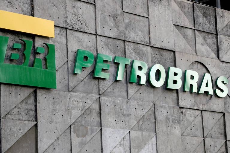 A imagem mostra o logotipo da Petrobras em uma parede de concreto. O logotipo é composto pelas letras 'BR' em verde, com um retângulo amarelo acima, e a palavra 'PETROBRAS' em letras grandes e verdes. A parede apresenta um padrão de triângulos e texturas de concreto.
