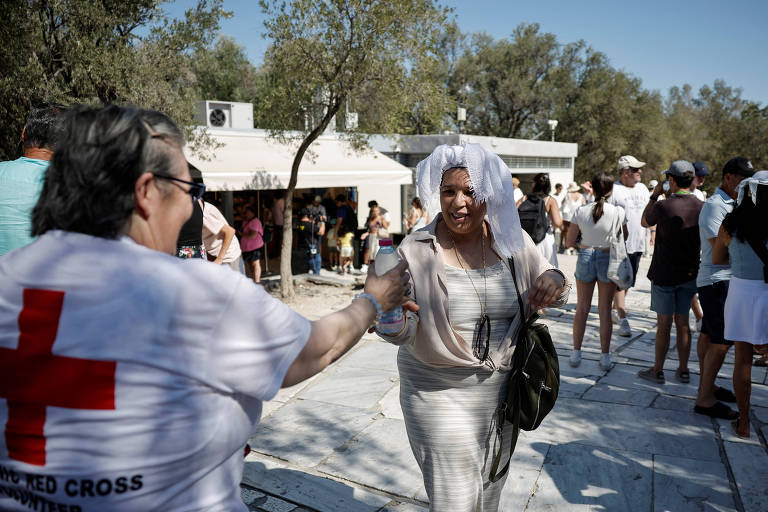 Mulher com camiseta com cruz vermelha estampada nas costas entrega garrafa plástica de água para outra mulher que passa com um pano amarrado na cabeça para proteger o rosto do sol