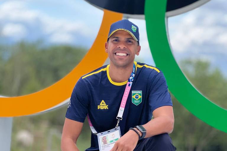 Um homem sorridente está agachado em frente aos anéis olímpicos coloridos. Ele usa uma camiseta azul com detalhes amarelos e um boné azul, ambos com o logo do Brasil. O homem também está usando calças escuras e tênis amarelos. Ele tem um crachá pendurado no pescoço, que parece ser de um evento olímpico. O fundo mostra um céu com nuvens e uma área verde.