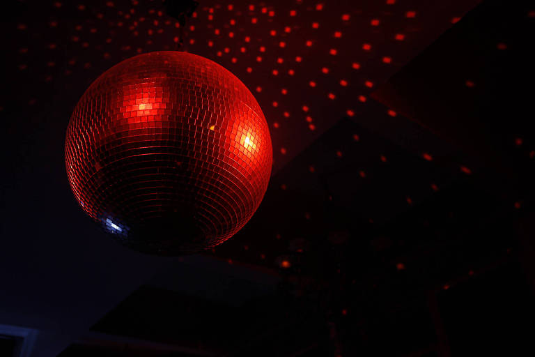 A imagem mostra uma bola de discoteca suspensa no teto, refletindo luzes vermelhas que criam um padrão de pontos no ambiente escuro ao redor.