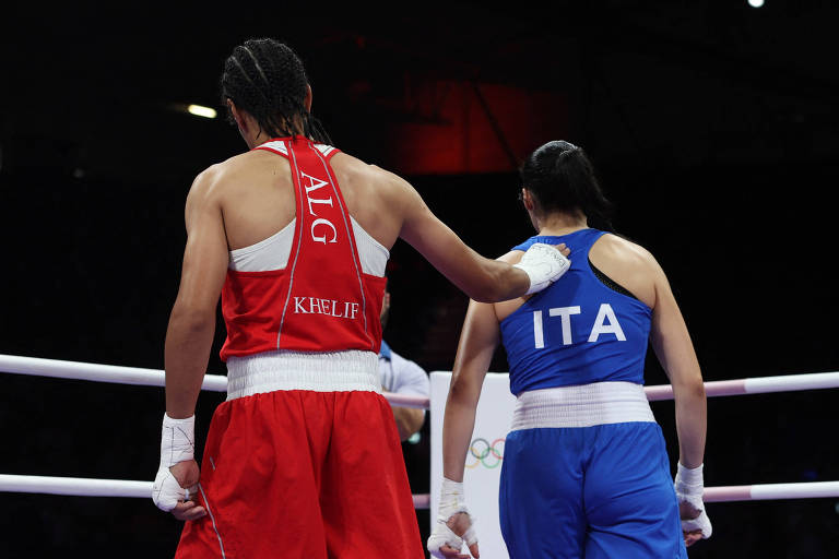 A imagem mostra duas boxeadoras em um ringue. A boxeadora à esquerda está vestindo um uniforme vermelho e a boxeadora à direita está vestindo um uniforme azul. Ambas estão de costas, com a boxeadora de vermelho colocando a mão no ombro da boxeadora de azul. O ambiente é iluminado, com um fundo desfocado que sugere um evento esportivo.