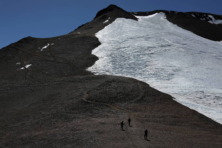 A imagem mostra uma paisagem montanhosa com um grande glaciar à direita. Três pessoas estão caminhando em direção ao glaciar, em um terreno rochoso e irregular. O céu está limpo e azul, e a luz do sol ilumina a cena, destacando a textura das rochas e a neve