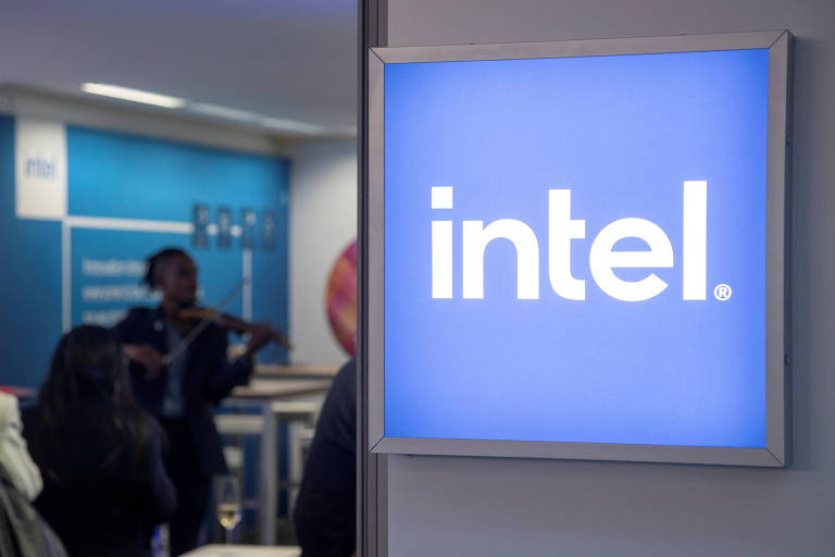 A imagem mostra um painel com o logotipo da Intel em um fundo azul. Ao fundo, há uma pessoa tocando violino, enquanto outras pessoas estão visíveis em primeiro plano, algumas com taças de bebida. O ambiente parece ser um evento ou uma conferência.
