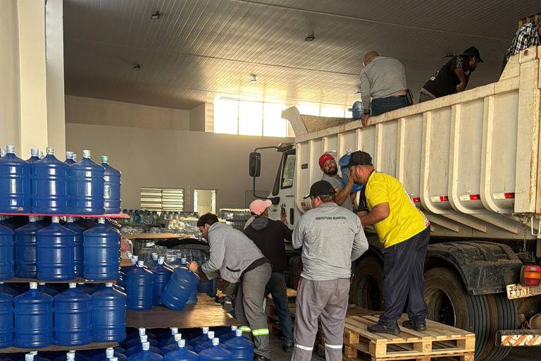 A imagem mostra um grupo de pessoas trabalhando em um armazém, carregando galões azuis de água. Alguns homens estão em cima de um caminhão, enquanto outros estão organizando os galões em paletes