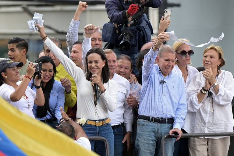 A imagem mostra um grupo de pessoas em um evento ao ar livre. No centro, uma mulher de cabelo longo e escuro segura um microfone e levanta o braço, enquanto outras pessoas ao seu redor também levantam os braços e seguram papéis. Há um homem ao seu lado, com cabelo grisalho, que também está levantando a mão. O fundo é composto por uma multidão e uma bandeira colorida visível na parte inferior da imagem.
