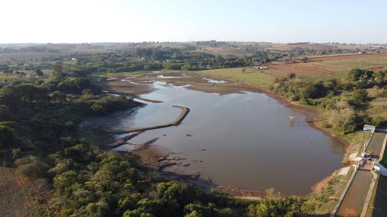 A imagem mostra uma vista aérea de uma lagoa com áreas de terra exposta ao redor. A água é de cor marrom e há vegetação densa nas margens. No fundo, pode-se ver uma paisagem rural com colinas e algumas casas