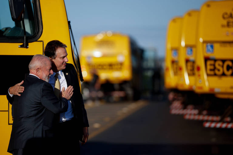 A imagem mostra Lula e Camilo Santana em um abraço próximo a um ônibus escolar amarelo. Ao fundo, há vários ônibus amarelos estacionados, com a luz do sol iluminando a cena