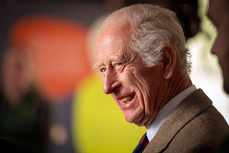 A imagem mostra Charles, um homem de cabelos brancos e pele clara, sorrindo levemente. Ele está usando um paletó e parece estar em um ambiente com fundo desfocado, onde há formas coloridas. O foco está em seu rosto, que expressa uma atitude amigável.