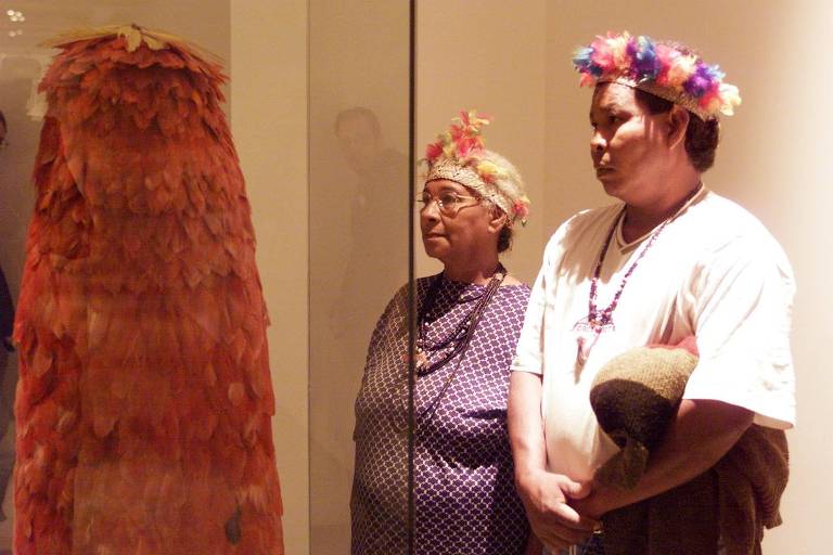 Um homem e uma mulher, ambos indígenas, observam um manto feito de penas vermelhas protegidos por um vidro. 
