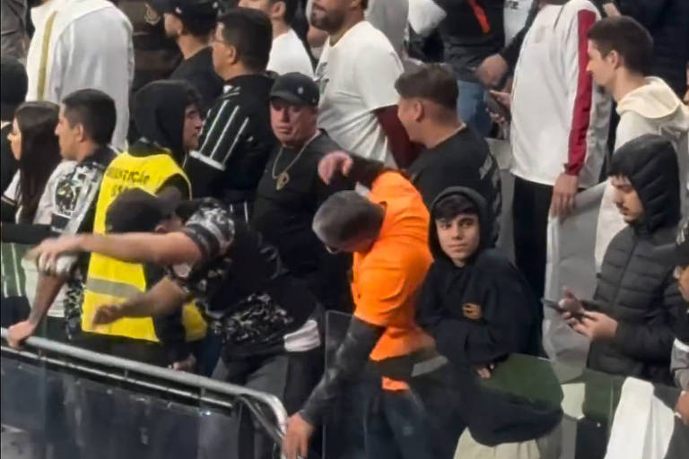 A imagem mostra uma multidão de torcedores em um evento esportivo. As pessoas estão vestidas com camisetas de diferentes cores, incluindo preto e branco, e algumas usam jaquetas. Há um torcedor em destaque, vestindo uma camiseta laranja, que simula gestos de natação junto com outro torcedor.