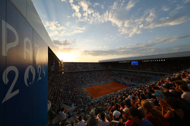 A imagem mostra um estádio de tênis durante o pôr do sol, com uma grande quantidade de espectadores nas arquibancadas. O céu está parcialmente nublado, com raios de sol visíveis. O piso da quadra é de terra batida, e há uma tela de exibição visível ao fundo.
