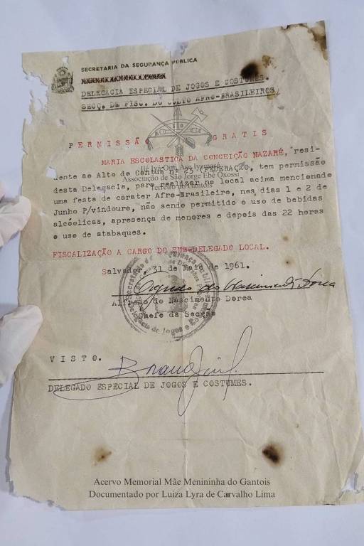 Documento oficial datado de 31 de maio de 1961, emitido pela Delegacia Especial de Jogos e Costumes da Secretaria da Segurança Pública. O documento concede permissão a Maria Escolástica da Conceição Nazaré para realizar uma festa de caráter Afro-Brasileiro nos dias 1 e 2 de junho, com restrições sobre o uso de bebidas alcoólicas e a presença de menores. O documento contém assinaturas e um carimbo oficial.