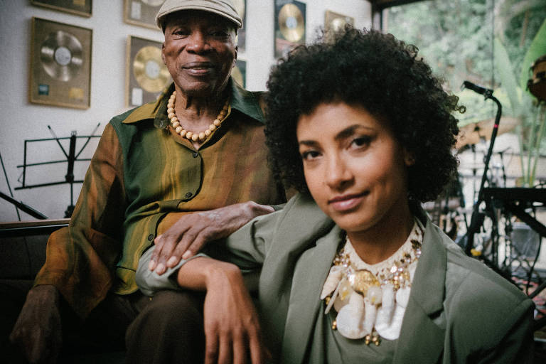 A imagem mostra um homem idoso e uma mulher jovem sentados juntos em um ambiente musical. O homem usa uma camisa de cores verde e marrom, um colar de contas e um boné. A mulher, com cabelo cacheado, veste um blazer cinza e um colar decorativo. Ao fundo, há prêmios de discos dourados nas paredes e instrumentos musicais visíveis.