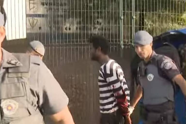 A imagem mostra um grupo de policiais em uma operação. Um homem, vestido com uma camisa listrada em preto e branco e calças escuras, está sendo levado sob custódia por um policial. O policial à direita está segurando o homem, enquanto outro policial, em primeiro plano, está parcialmente visível. Ao fundo, há uma cerca e uma estrutura urbana