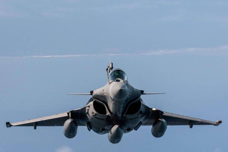 Avião militar cinza é visto de frente, com três tanques embaixo das asas, em um céu azul