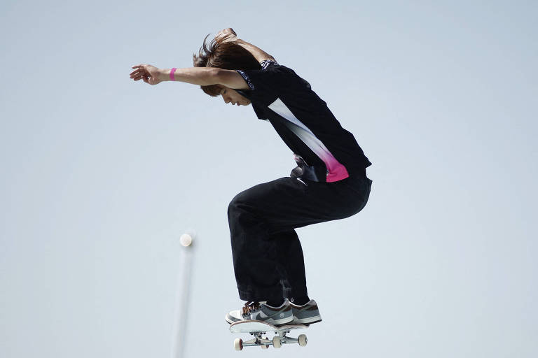 Um skatista está realizando um salto sobre o skate, com os pés elevados e o corpo inclinado para frente. Ele usa uma camiseta preta com detalhes em rosa e calças escuras. O fundo é claro, sugerindo um dia ensolarado.