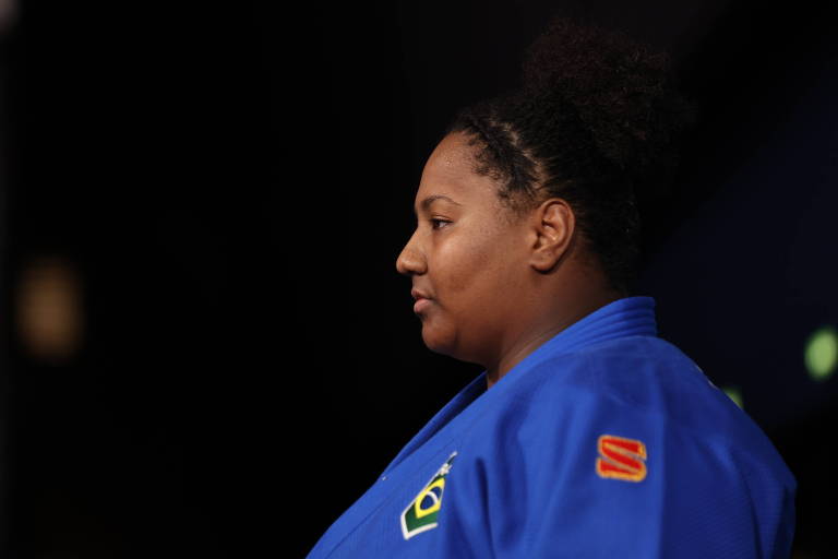 A imagem mostra uma atleta de judô em perfil, vestindo um judogi azul. Ela tem cabelo crespo preso em um coque e está em um ambiente com fundo escuro. O judogi possui um emblema do Brasil no lado esquerdo do peito e uma marca na manga direita.
