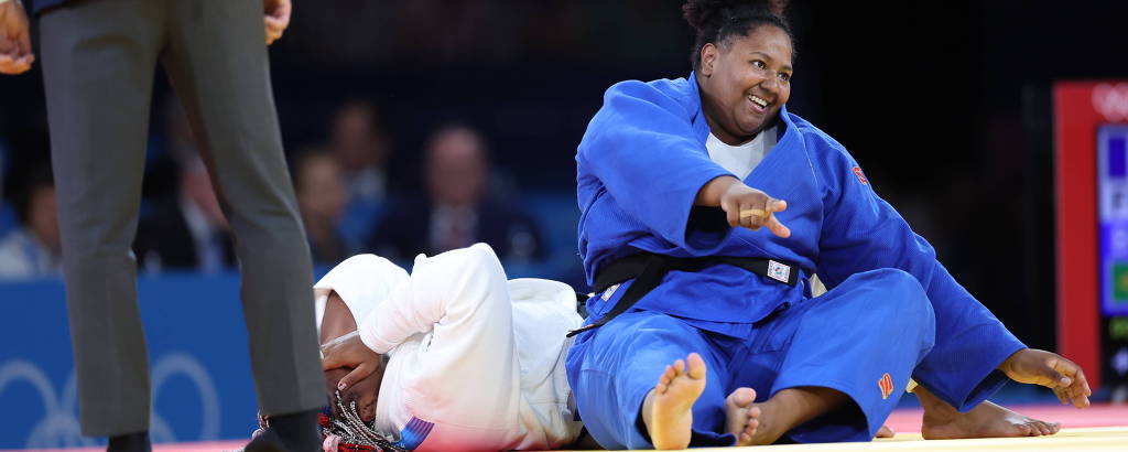 A imagem mostra duas competidoras de judô em uma luta. Uma atleta está vestido com um quimono azul, sentada no tatame, enquanto a outra, vestido de branco, está deitada no chão. Um juiz, parcialmente visível, está em pé ao fundo, observando a ação.