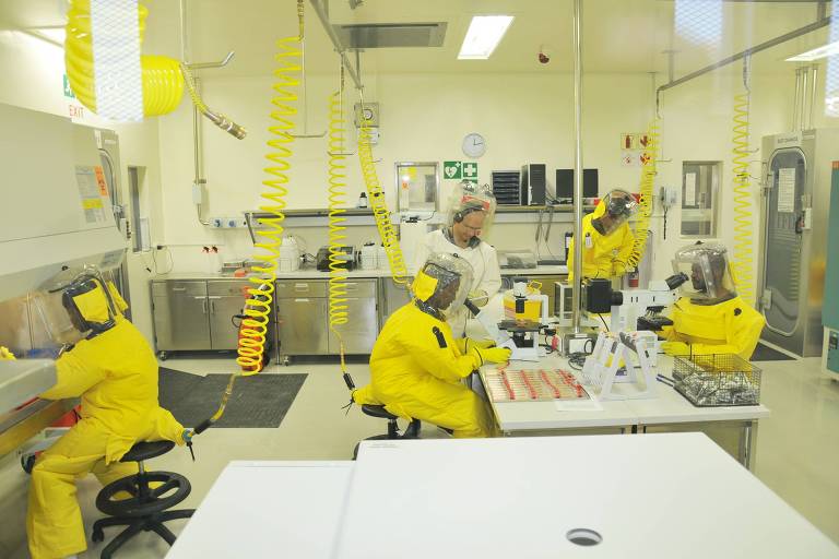A imagem mostra um laboratório com várias pessoas usando trajes de proteção amarelos. Há quatro indivíduos visíveis, dois deles sentados em mesas de trabalho, um em pé e outro em uma estação de trabalho. O ambiente é limpo e bem iluminado, com equipamentos de laboratório ao fundo e paredes brancas
