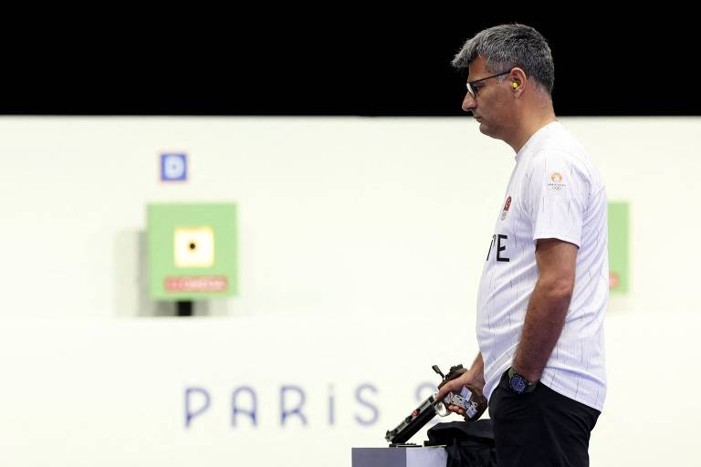 Um homem está em pé, segurando uma arma, com uma expressão séria. Ao fundo, há alvos de tiro e a palavra 'PARIS' visível. O homem usa uma camiseta branca e parece estar concentrado na competição.
