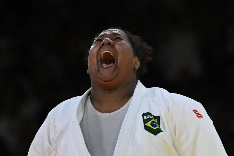 A imagem mostra um atleta de judô, vestindo um quimono branco com um emblema verde e amarelo, expressando uma forte emoção ao gritar. O fundo é desfocado, sugerindo um ambiente de competição.
