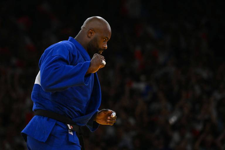 Um atleta de judô, vestido com um quimono azul, está em movimento, com um gesto de comemoração ou determinação. O fundo mostra uma multidão, sugerindo um evento esportivo. O foco está no atleta, que parece concentrado e energizado.

