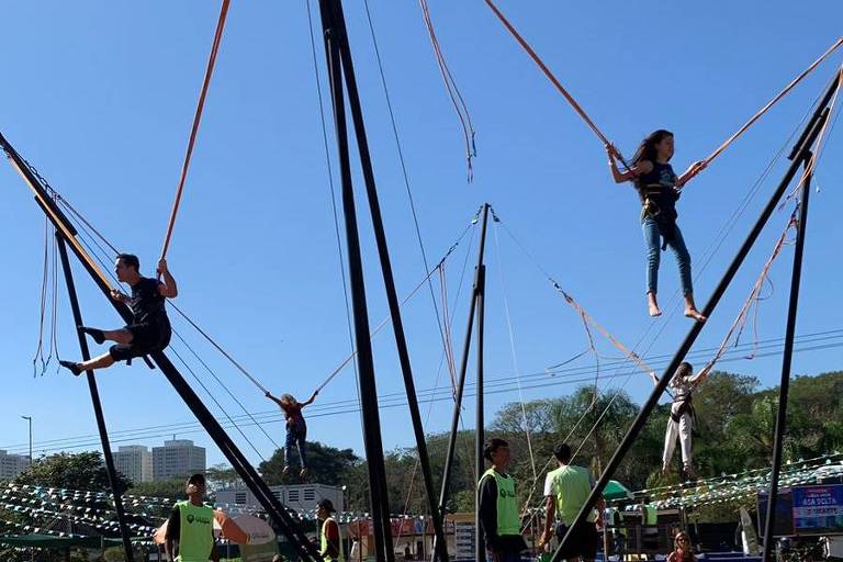 A  imagem mostra uma estrutura de trampolim com várias pessoas se divertindo. Há três crianças pulando em trampolins, presas a cordas que se estendem para cima. Algumas pessoas estão em pé ao redor da estrutura, vestindo coletes verdes. O céu está claro e azul, e ao fundo, pode-se ver uma área de parque com árvores e outras atrações.