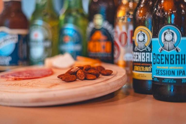 A imagem mostra uma mesa com várias garrafas de cerveja dispostas ao fundo, incluindo uma garrafa da Eisenbahn. Em primeiro plano, há uma tábua de madeira com fatias de salame, amêndoas e um pedaço de queijo. A mesa tem um ambiente descontraído, ideal para um encontro social.